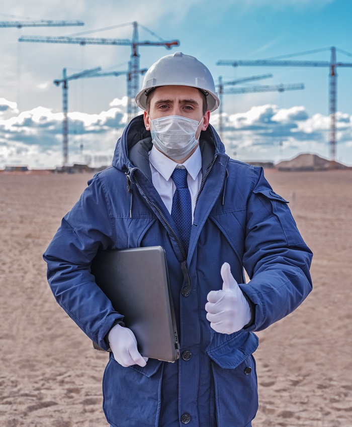 a civil engineer with a laptop, wearing a protective medical mask, gloves and a white helmet against the background of the construction start.